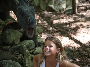 Ein Mädchen mit langen Haaren lächelt fröhlich, während ein Dinosauriermodell hinter ihr steht und den Mund geöffnet hat.