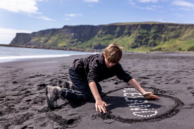 Ein Junge kniet am schwarzen Sandstrand und gestaltet mit Steinen eine kreative Form im Sand.