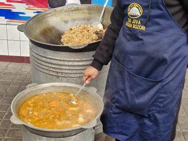 Ein Koch in einem blauen Schürze rührt in einem großen Topf mit dampfendem Eintopf, während eine Frau im Hintergrund zusieht.