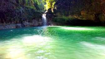 Ein klarer, türkisfarbener Wasserfall fließt sanft in einen ruhigen, grünen Pool, umgeben von üppigem, grünem Laub.