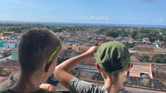 Zwei Jungen stehen an einem Aussichtspunkt und blicken auf die bunten Dächer einer Stadt und das Meer in der Ferne.