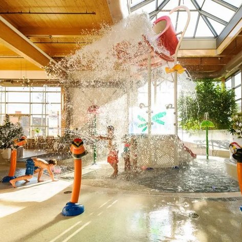 Ein lebhafter Wasserpark mit spritzenden Kindern, die in einem hellen, sonnendurchfluteten Raum spielen.