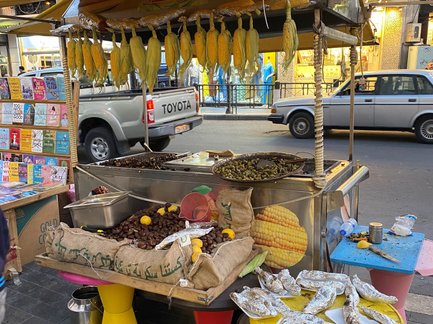 Ein Straßenstand bietet frisches Obst und gegrillten Fisch an, umgeben von bunten Tischen und einer belebten Straße.