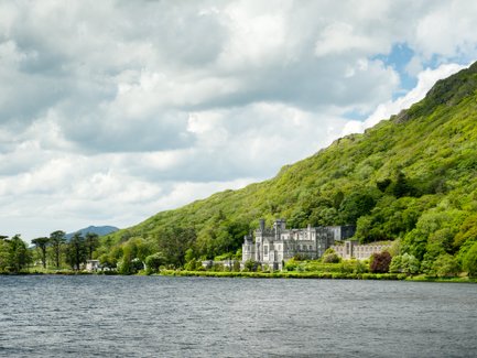 Ein majestätisches Schloss liegt am Ufer eines ruhigen Sees, umgeben von üppigem, grünem Wald und sanften Hügeln.