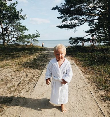 Ein kleines Kind in einem weißen Bademantel läuft barfuß auf einem Weg zum Strand, umgeben von Bäumen und Wasser.