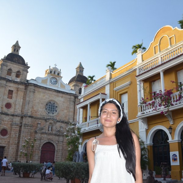 Eine Frau mit langen, schwarzen Haaren steht vor der Kirche San Pedro Claver, umgeben von bunten Gebäuden in Cartagena.