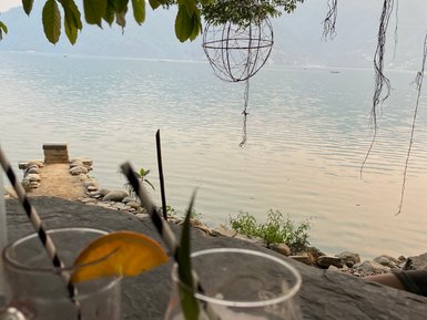 Zwei Cocktails mit bunten Strohhalmen stehen auf einem Tisch, während der ruhige See im Hintergrund glitzert.