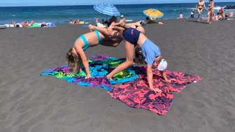 Zwei Mädchen machen akrobatische Übungen auf bunten Handtüchern am Strand, umgeben von anderen Sonnenanbetern.