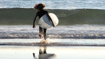 Ein Surfer mit langen Haaren läuft ins Wasser, während die Wellen sanft an den Strand rollen.