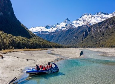 Ein Jetboot mit mehreren Passagieren gleitet über den klaren Fluss, umgeben von majestätischen Bergen und strahlend blauem Himmel.