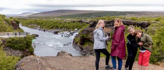 Eine Gruppe von vier Personen steht am Rand eines Wasserfalls, umgeben von grüner Landschaft und einem fließenden Fluss.