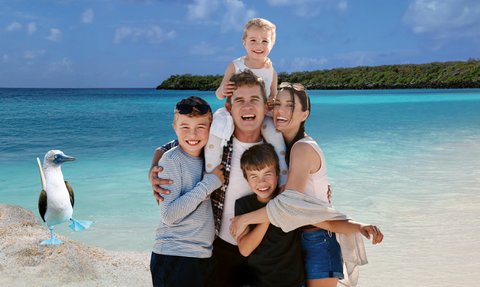 Eine fröhliche Familie steht am Strand, umarmt sich und lacht, während im Hintergrund das klare Meer und der Himmel leuchten.