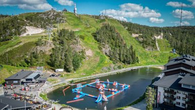 Ein lebhaftes Wasserpark-Set mit bunten Attraktionen auf einem klaren See, umgeben von grünen Hügeln und blauen Himmel.