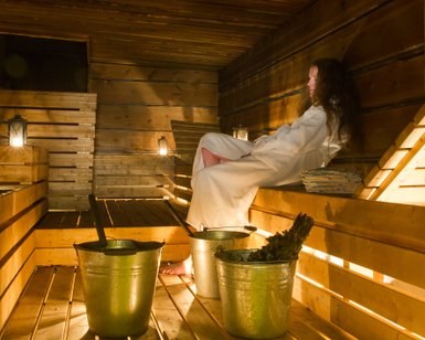 In einer warmen Sauna sitzt eine Person in einem weißen Bademantel entspannt auf einer Holzbank, umgeben von Eimern.