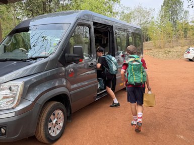 Zwei Kinder steigen in einen grauen Minibus ein, während sie Rucksäcke tragen und ein Kind eine Papiertüte hält.