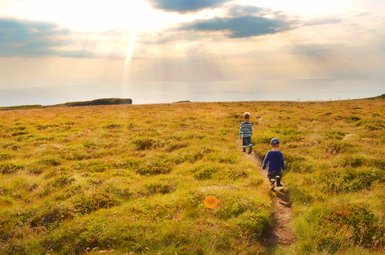 Zwei Kinder wandern auf einem schmalen Pfad durch eine grüne Wiese, während die Sonne über dem Meer aufgeht.
