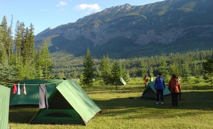 Zwei Personen stehen auf einem Campingplatz mit grünen Zelten, umgeben von hohen Bäumen und majestätischen Bergen im Hintergrund.