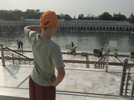 Ein Junge mit einem orangefarbenen Turban steht am Ufer eines ruhigen Gewässers und blickt auf die Menschenmenge.
