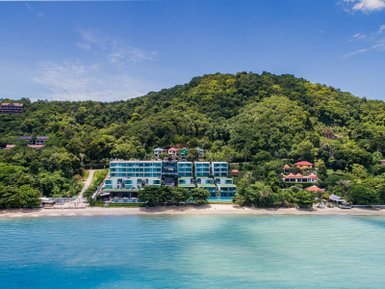 Ein modernes Hotel am Strand, umgeben von üppigem Grün und sanften Hügeln, unter einem strahlend blauen Himmel.