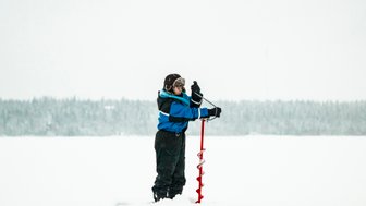 Ein Kind in blauer Winterkleidung bohrt mit einer roten Eisbohrmaschine in eine schneebedeckte Landschaft.