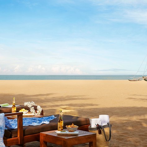 Ein entspannender Strandbereich mit Liegen, einem Tisch mit Snacks und Getränken sowie einem Segelboot im Hintergrund.