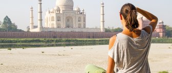 Eine Frau sitzt am Ufer und betrachtet das majestätische Taj Mahal, umgeben von einer ruhigen Landschaft.