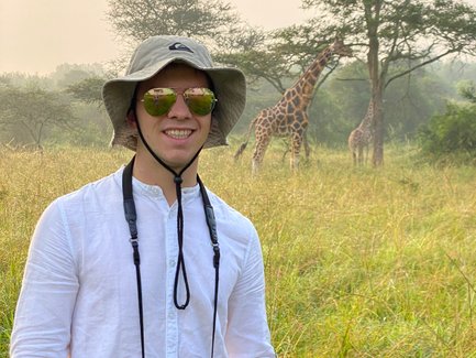 Ein junger Mann mit Sonnenbrille und Hut steht in einer grünen Savanne, während Giraffen im Hintergrund grasen.