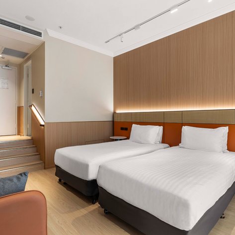 Ein modernes Hotelzimmer in Sydney mit zwei Einzelbetten, einem gemütlichen Sessel und einer eleganten Holzverkleidung.