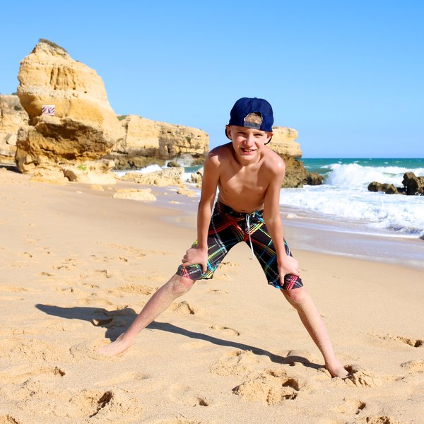 Ein fröhlicher Junge steht am Strand, mit weit gespreizten Beinen und einem breiten Lächeln, während die Wellen sanft an den Strand rollen.