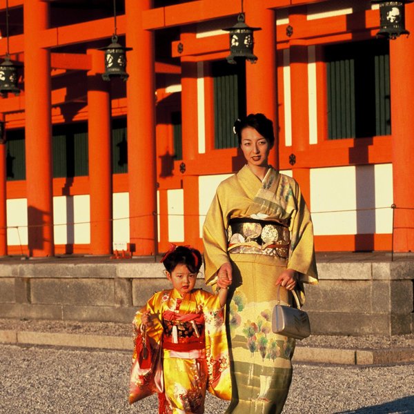 Eine Frau in einem eleganten Kimono und ein kleines Mädchen in einem bunten Kimono spazieren gemeinsam vor einem historischen Gebäude.