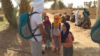 Eine Gruppe von Kindern in bunten Tüchern hört einem Erwachsenen mit Rucksack zu, während Dromedare im Hintergrund warten.