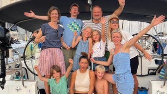 Eine fröhliche Gruppe von Erwachsenen und Kindern posiert auf einer Segelyacht, umgeben von Masten und einem schönen Himmel.