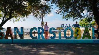 Bunte Buchstaben bilden den Schriftzug 'SAN CRISTOBAL' vor einer malerischen Küstenlandschaft auf den Galapagos-Inseln.