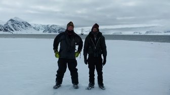 Zwei Männer stehen auf einer schneebedeckten Fläche, umgeben von Bergen und einem Fjord im Hintergrund.