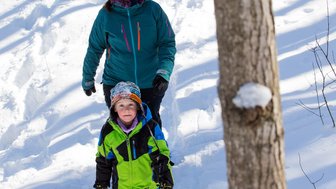 Eine Frau und ein Kind wandern fröhlich im Schnee, umgeben von schneebedeckten Bäumen und einem klaren blauen Himmel.