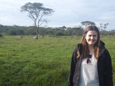 Eine Frau steht lächelnd in einer grünen Landschaft, während Zebras im Hintergrund grasen und ein Baum im Vordergrund sichtbar ist.