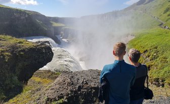 Zwei Personen stehen am Rand des Gullfoss-Wasserfalls und beobachten die tosenden Wassermassen, umgeben von grüner Landschaft.