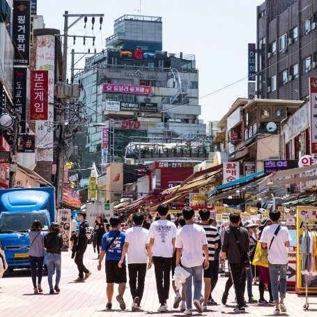 Eine belebte Einkaufsstraße in Hongdae, gesäumt von Geschäften und bunten Schildern, mit vielen Passanten im Sonnenlicht.