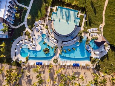 Ein atemberaubender Blick auf einen Poolbereich mit mehreren Schwimmbecken und Liegen, umgeben von Palmen und grünem Gras.