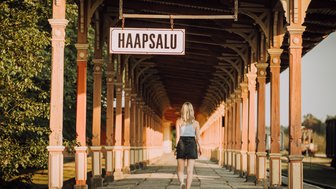 Eine Frau mit blonden Haaren geht auf einem historischen Bahnhofssteig in Haapsalu, umgeben von bunten Säulen.