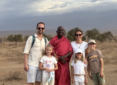 Eine Familie steht in der Savanne, umgeben von Bäumen, mit einem Maasai-Krieger in traditioneller Kleidung.
