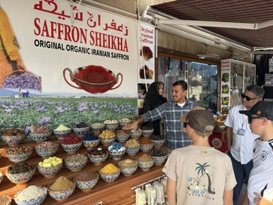 Ein Verkäufer zeigt eine Auswahl an Gewürzen und Saffran in bunten Schalen, während zwei Kinder und ein Erwachsener aufmerksam zuschauen.