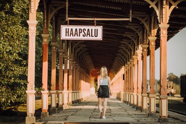 Eine Frau mit blonden Haaren geht auf einem historischen Bahnhofssteig in Haapsalu, umgeben von bunten Säulen.