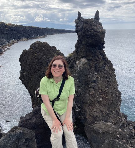 Eine Frau sitzt auf einem großen, dunklen Felsen am Meer, lächelnd und in einem hellgrünen Hemd gekleidet.