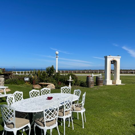 Ein einladender Garten mit einem großen Tisch, umgeben von weißen Stühlen, bietet einen Blick auf das Meer und den blauen Himmel.