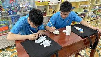 Zwei Jungen in blauen T-Shirts malen mit Pinseln auf schwarzen Stofftaschen, umgeben von bunten Bastelmaterialien.