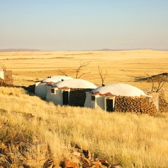 Drei runde, strohgedeckte Hütten stehen in einer weiten, goldenen Graslandschaft unter einem klaren Himmel.
