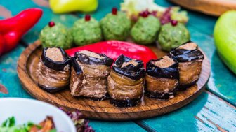 Ein Holzbrett präsentiert köstliche Pkhali-Röllchen aus Auberginen, umgeben von buntem Gemüse und frischen Kräutern.