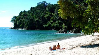 Ein sonniger Strand mit feinem Sand, umgeben von üppigem Grün und klarem Wasser, wo Menschen entspannen und schwimmen.