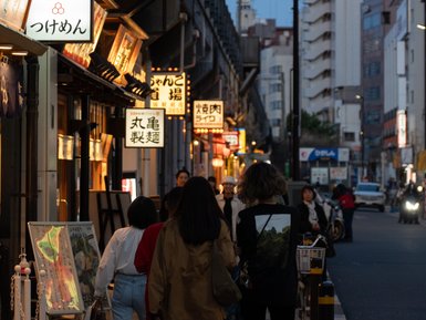 Eine belebte Straße in Tokio, gesäumt von Restaurants mit leuchtenden Schildern, während die Dämmerung einsetzt.